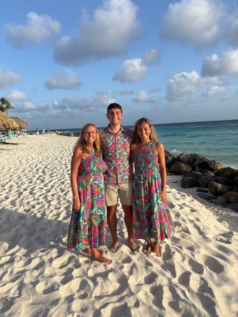 Three people in matching bright tropical print outfits smiling on a white sand beach with blue water behind them